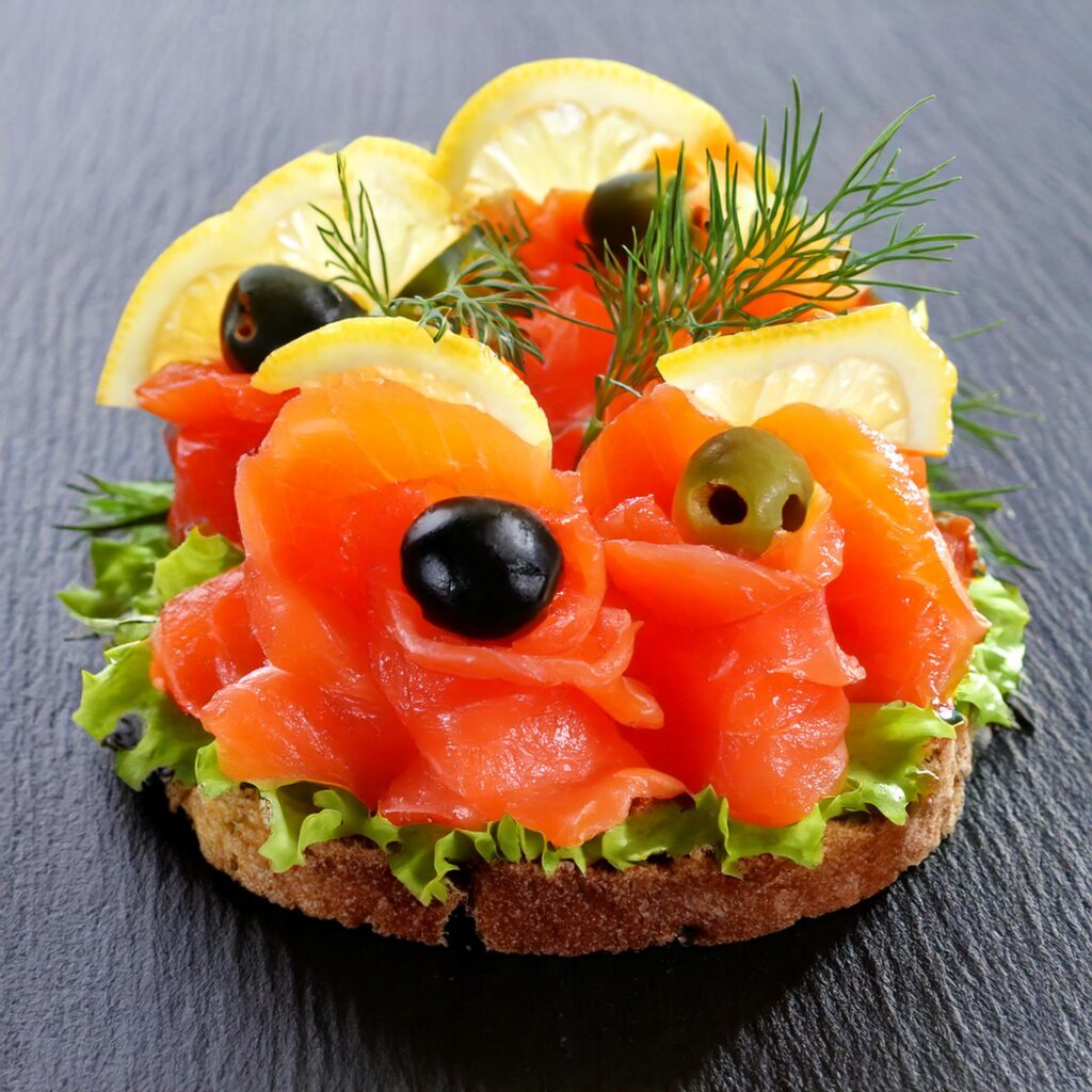 Smoked salmon appetizer on a slice of bread, garnished with fresh dill, black and green olives, lemon slices, and lettuce, presented elegantly on a slate platter.